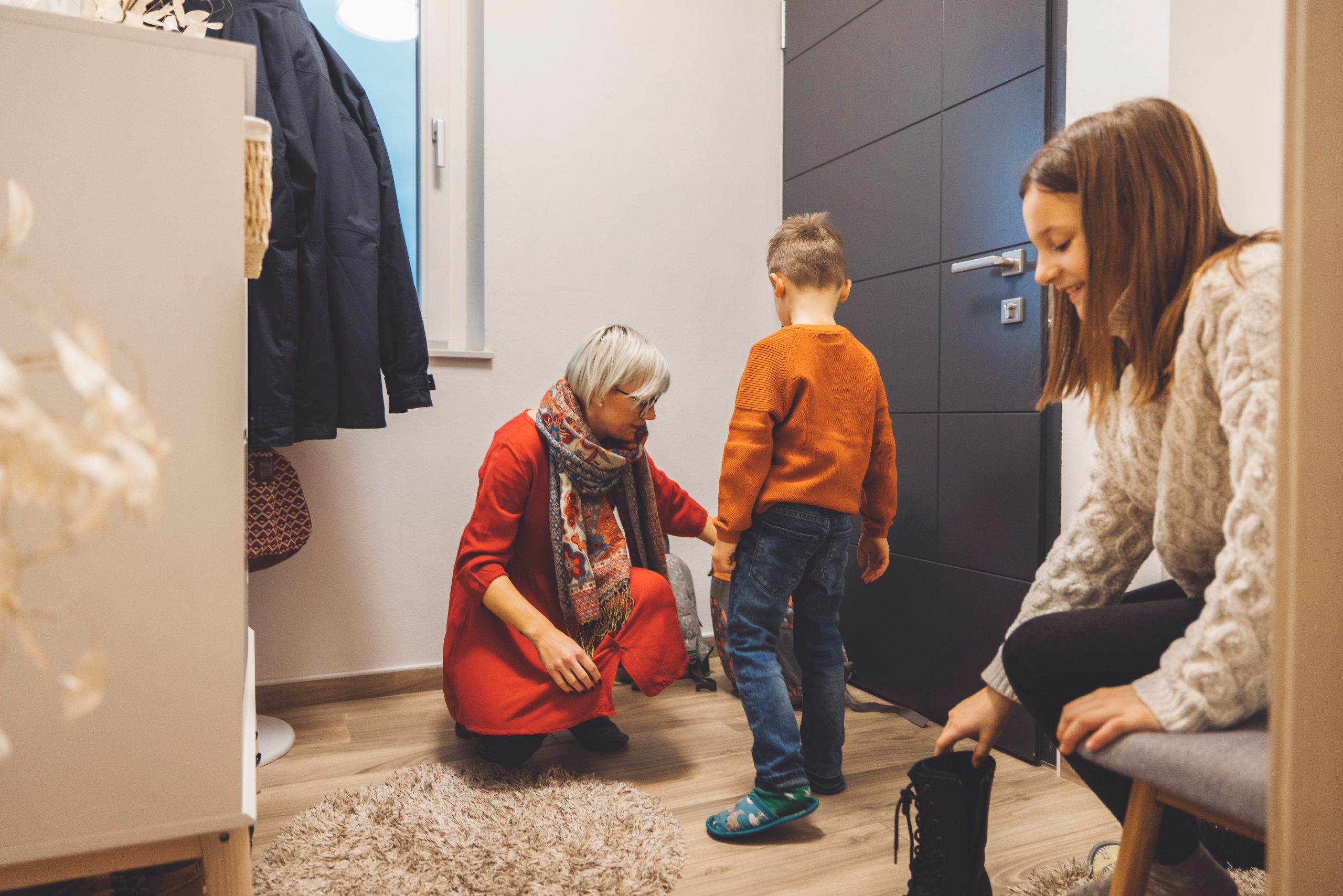 A woman in a red dress helps a young boy with his shoes in a cozy hallway. Another woman sits nearby, putting on her boots. Mother coming home from work, children hugging her with warm welcome. Uma mulher com um vestido vermelho ajuda um menino a calçar os sapatos em um corredor aconchegante. Outra mulher está sentada perto de você, calçando suas botas. Mãe chegando em casa do trabalho, com os filhos abraçando-a com calorosas boas-vindas.